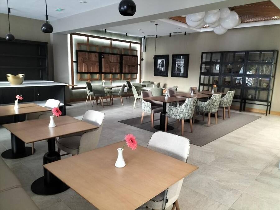 A modern dining area with multiple tables and chairs, a kitchen area with a black cabinet and a large window, and a wall with framed pictures.