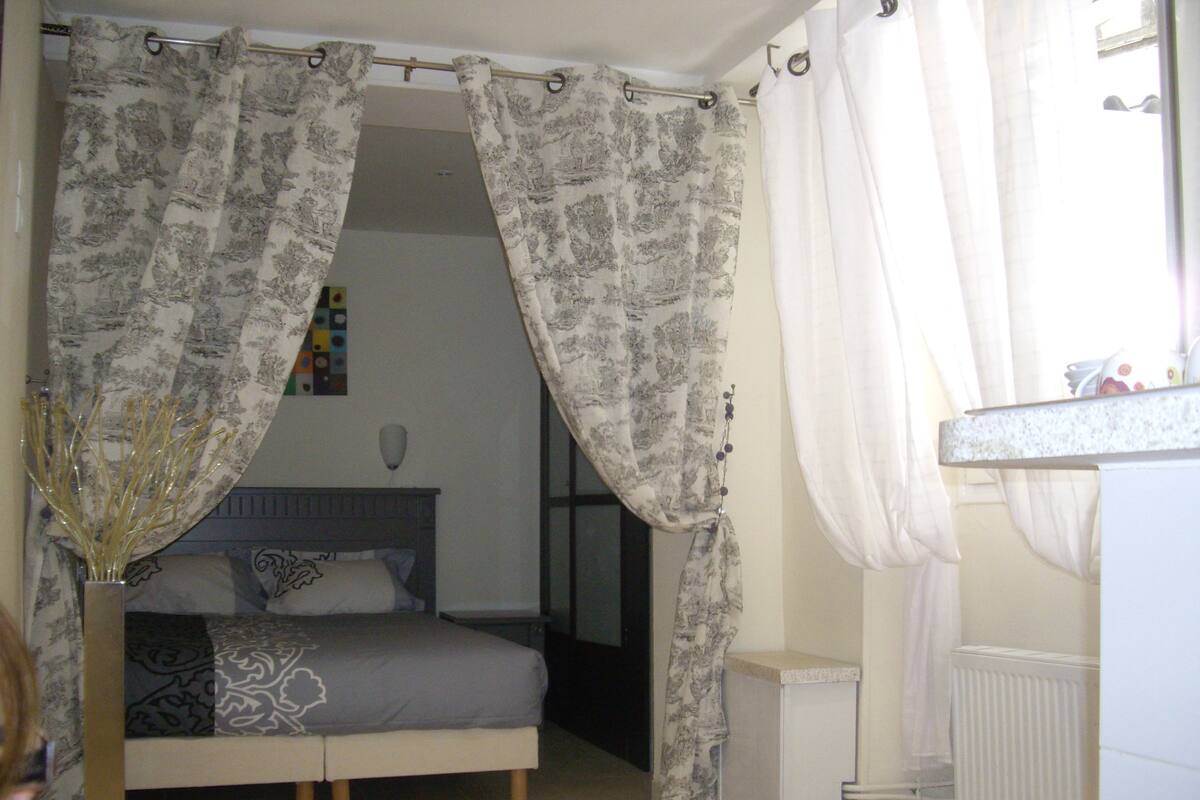 Quarto com cama, mesa de cabeceira, radiador e janela com cortinas.