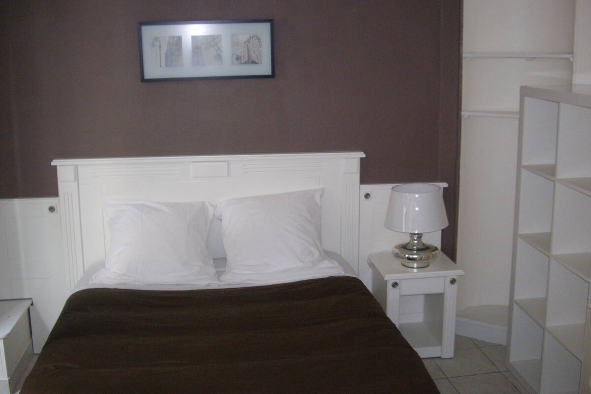 Quarto com cama branca, mesa de cabeceira com abajur e quadro na parede.