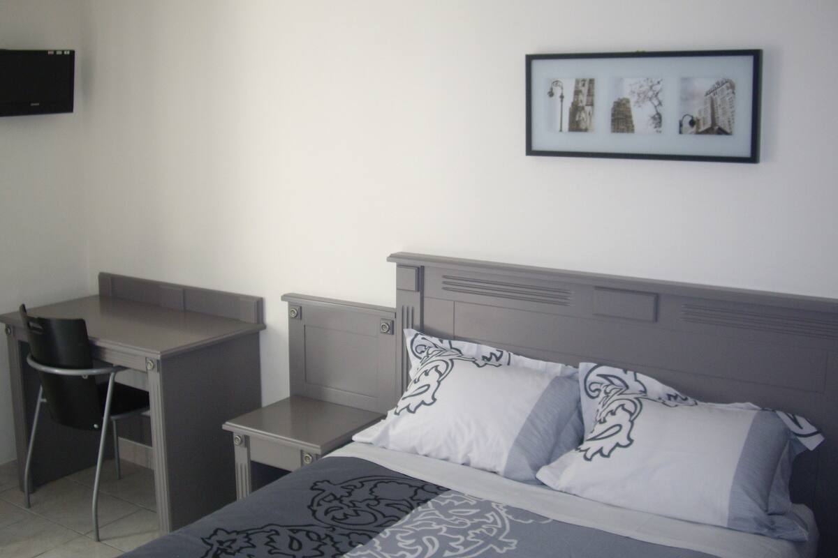 Quarto de hotel com uma cama, uma escrivaninha e uma cadeira. Há um quadro na parede.