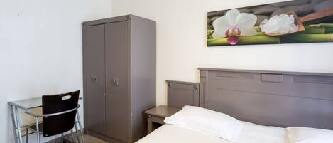 A hotel room with a bed, a desk with a chair, a wardrobe, and a painting of flowers on the wall.