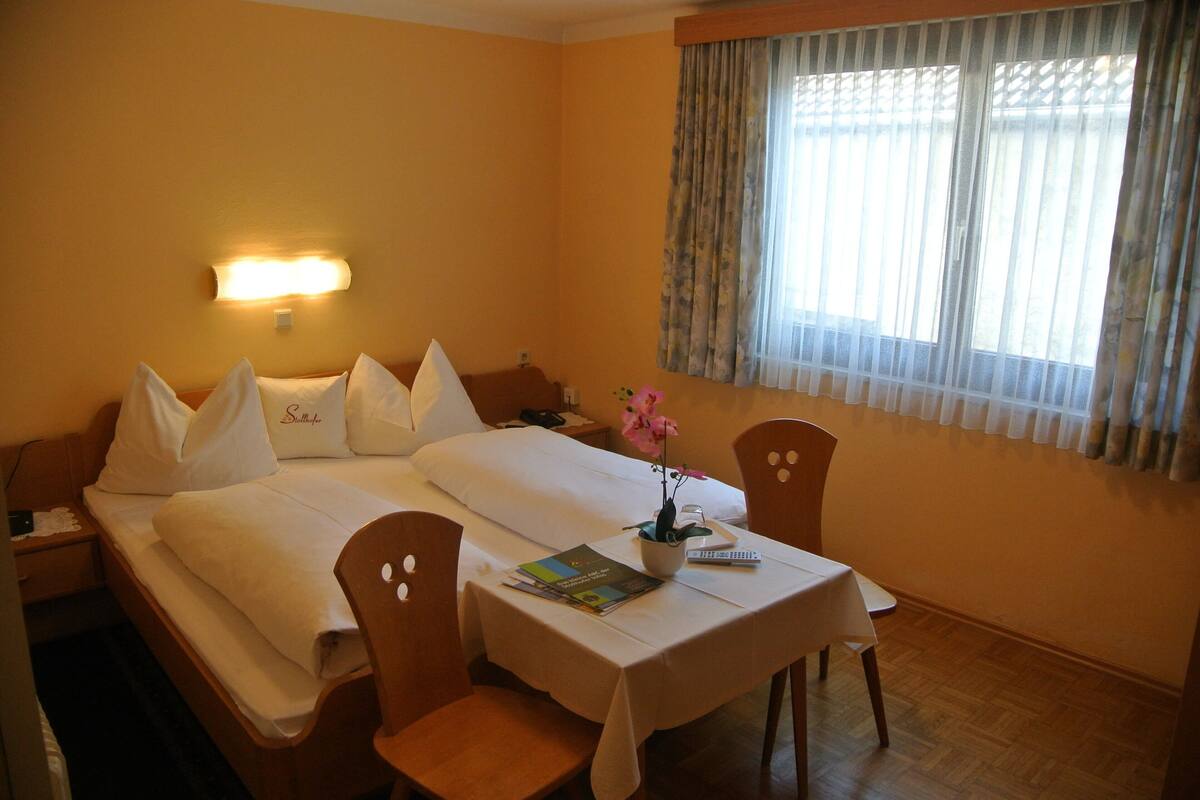 Habitación de hotel con cama doble, mesita de noche con lámpara, una pequeña mesa con una silla y una ventana con cortinas.