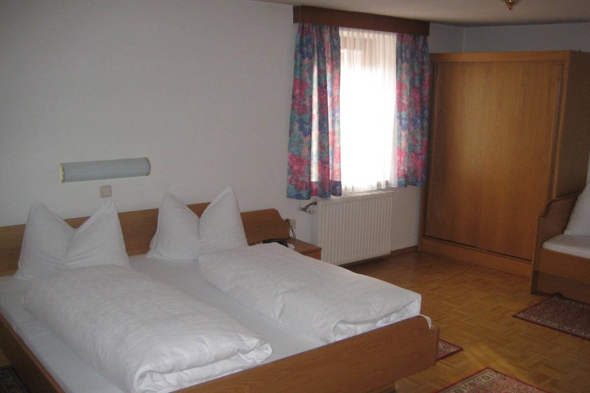 Un dormitorio con una cama de madera, ropa de cama blanca, un radiador, una ventana con cortinas florales y un armario de madera.