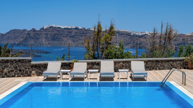 A swimming pool with lounge chairs, a stone wall, and a view of mountains and water. - Sigal Villa (Santorini)