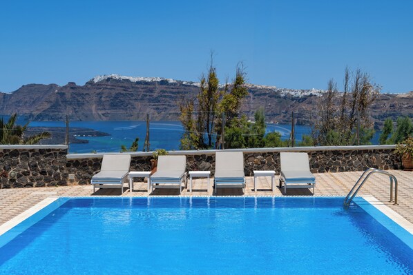 A swimming pool with lounge chairs, a stone wall, and a view of mountains and water. - Sigal Villa (Santorini)