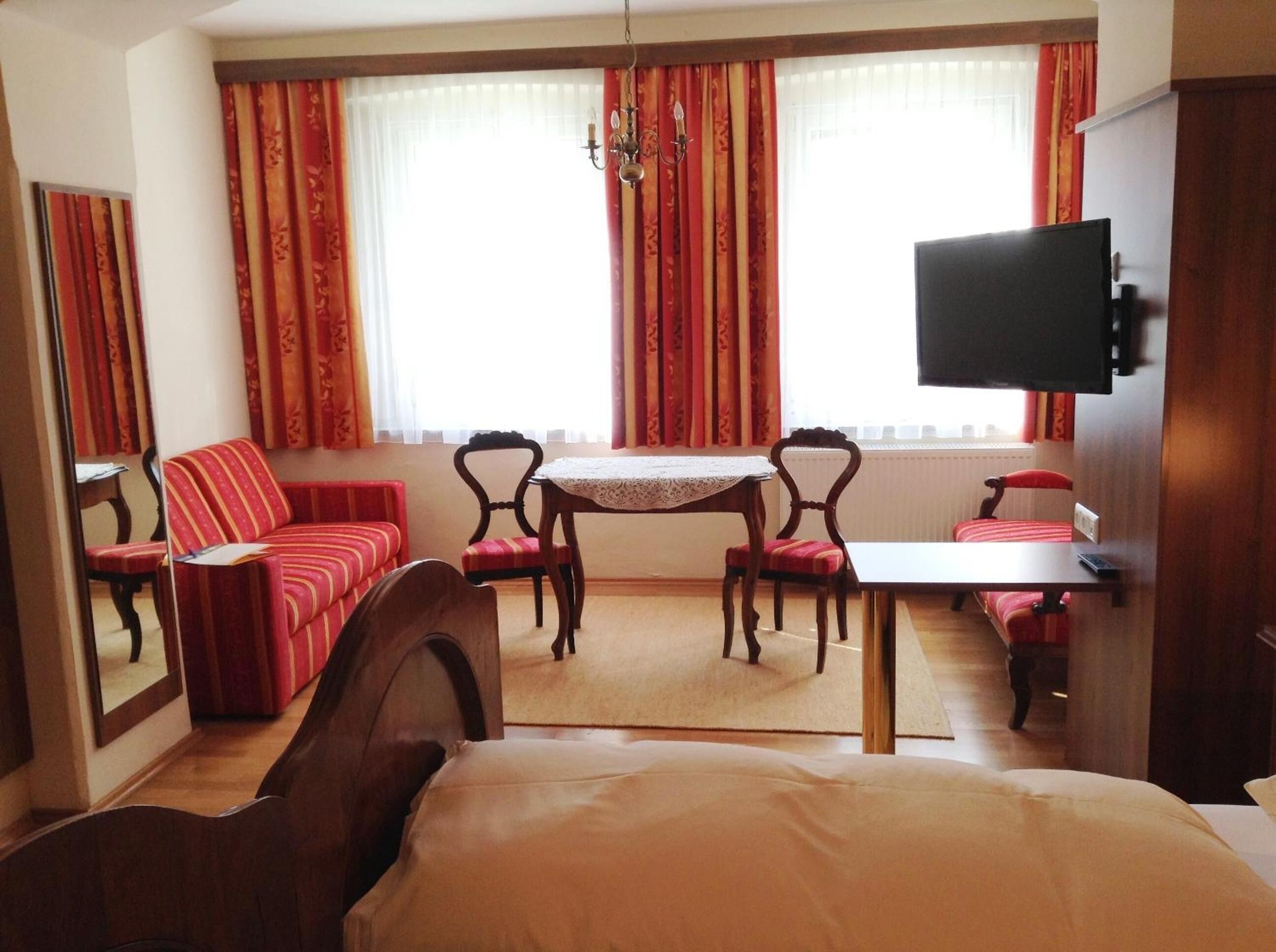 A hotel room with a bed, a sofa, a dining table with chairs, a television, and large windows with curtains.