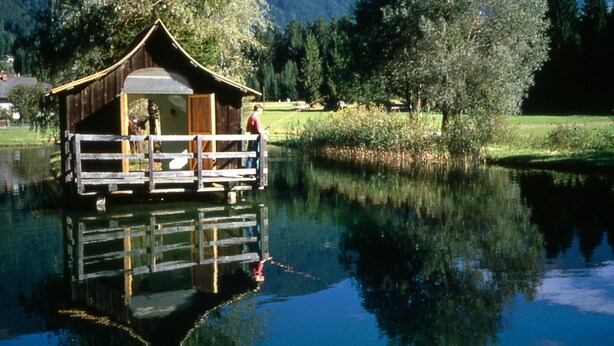 Una baita di legno con un balcone su un lago, circondata da alberi e montagne.