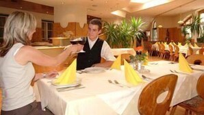 Un'ambientazione ristorante con tavoli apparecchiati, un cameriere che serve una bevanda e una sala da pranzo con sedie e tavoli.