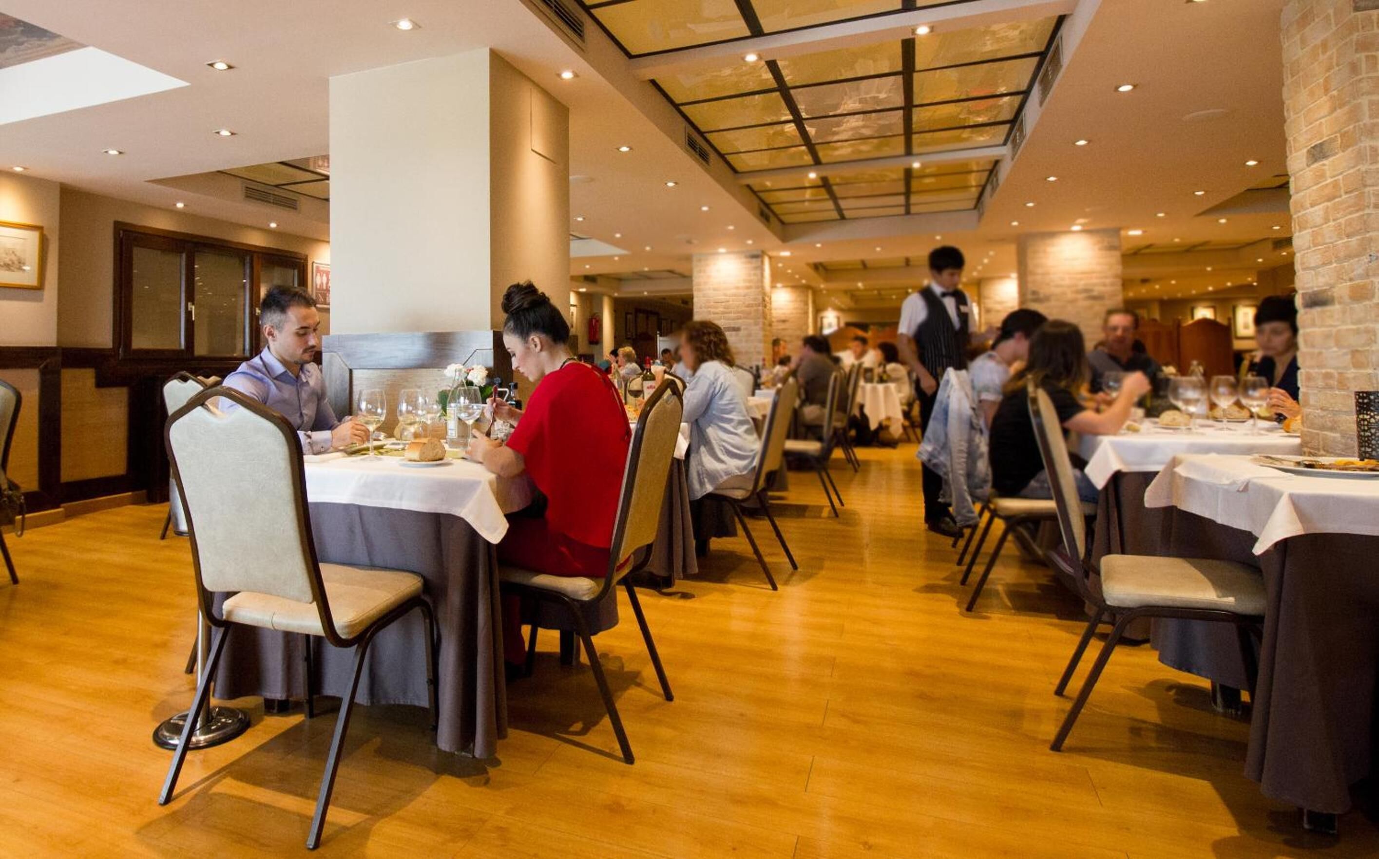 A restaurant with multiple tables, wooden flooring, and a high ceiling with skylights.