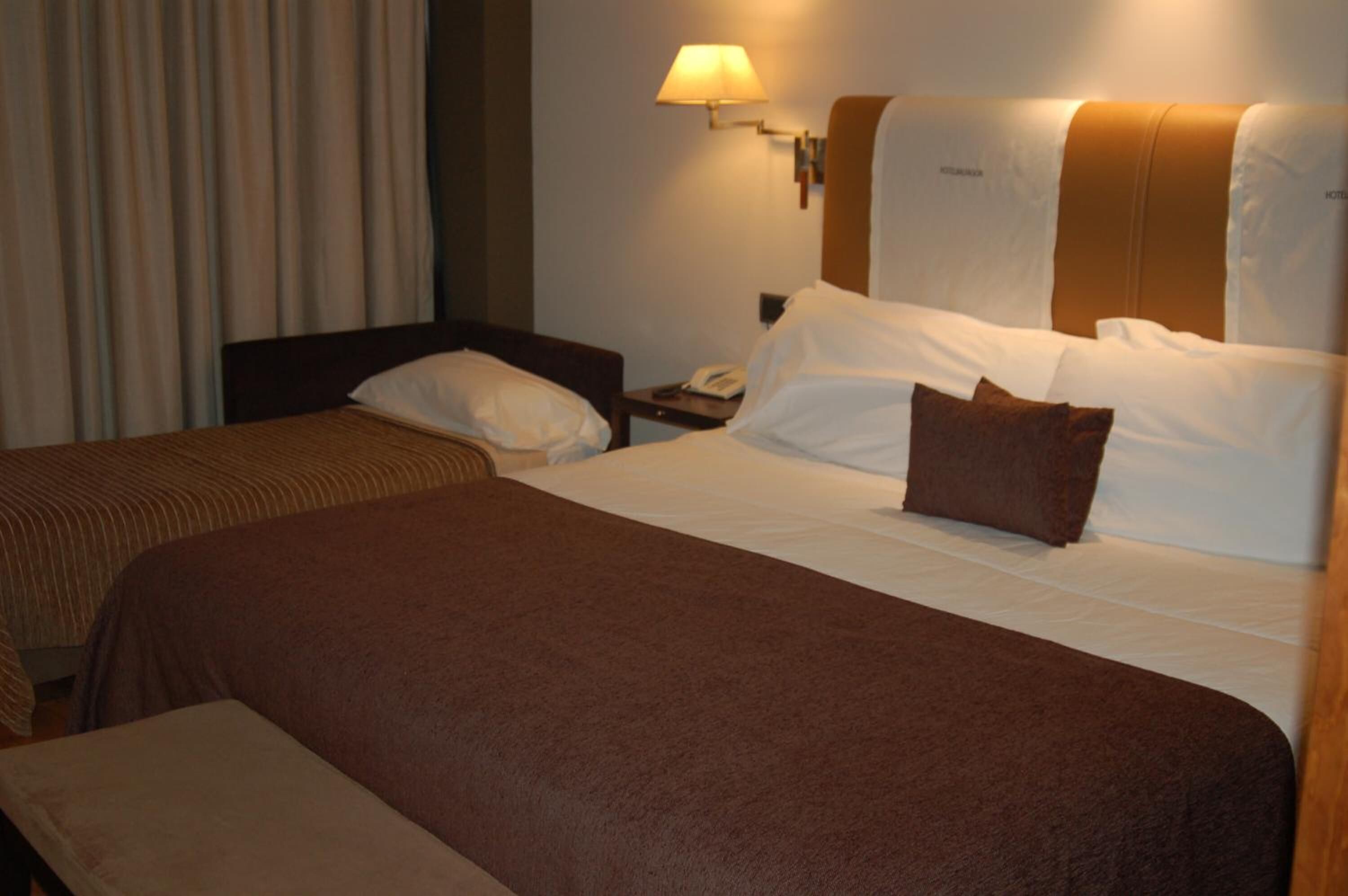 Een hotelkamer met een groot bed, een nachtkastje met een lamp en een raam met gordijnen.