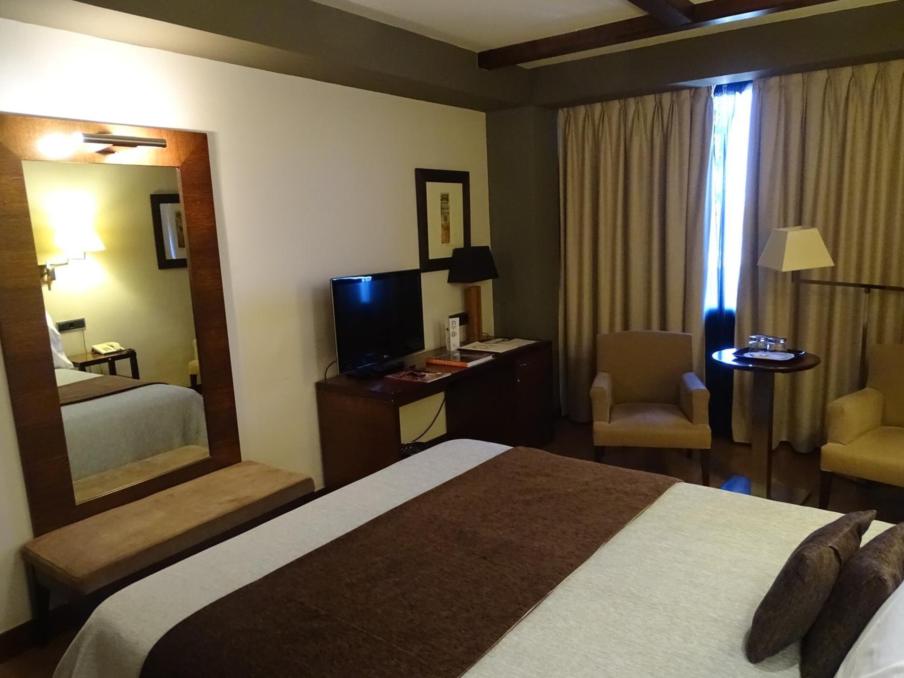 Habitación de hotel con una cama, un escritorio con computadora, una silla, una mesita y una ventana con cortinas.