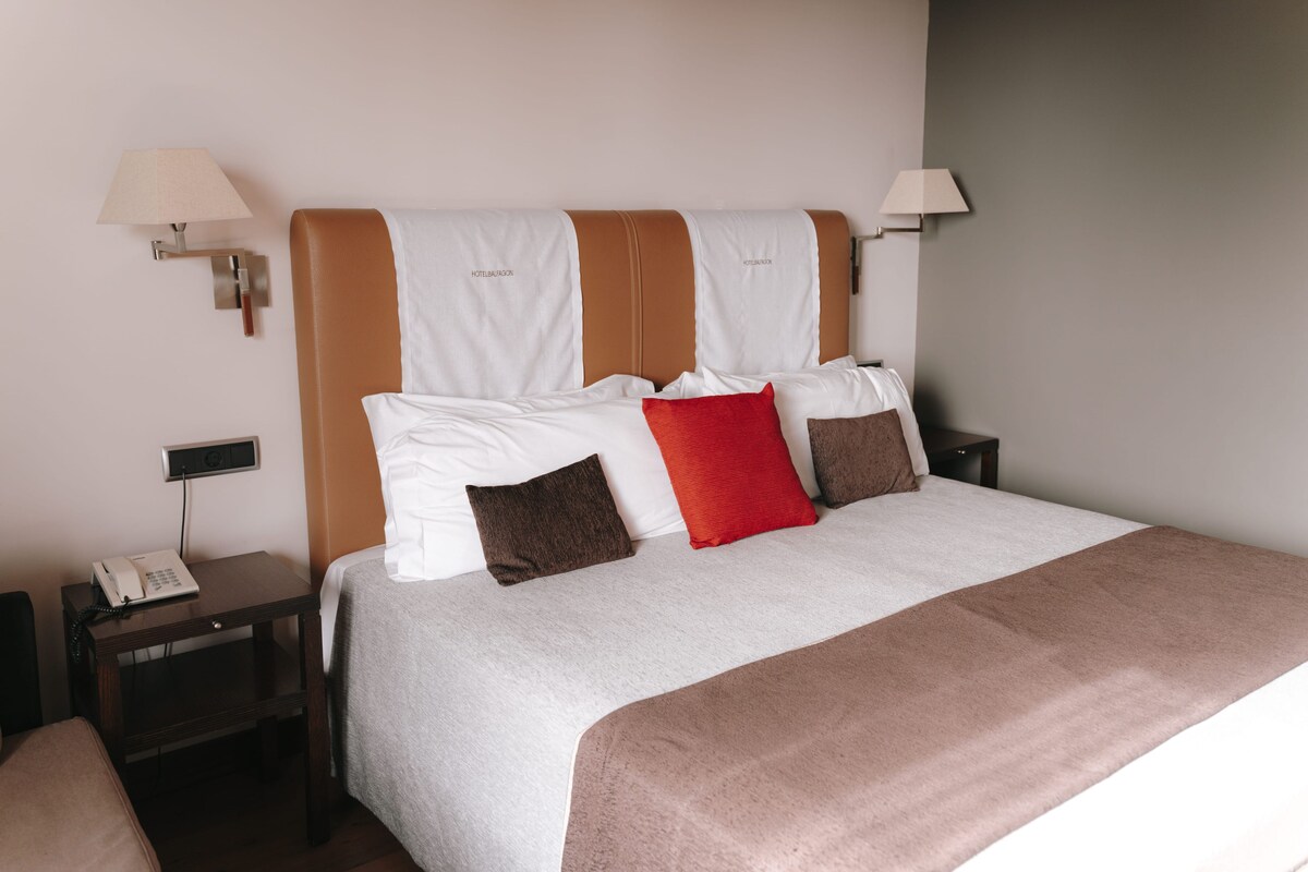 Una cama bien hecha con un cabecero marrón, almohadas blancas y marrones, y una colcha blanca. Hay dos mesitas de noche, una con una lámpara y la otra con un teléfono.