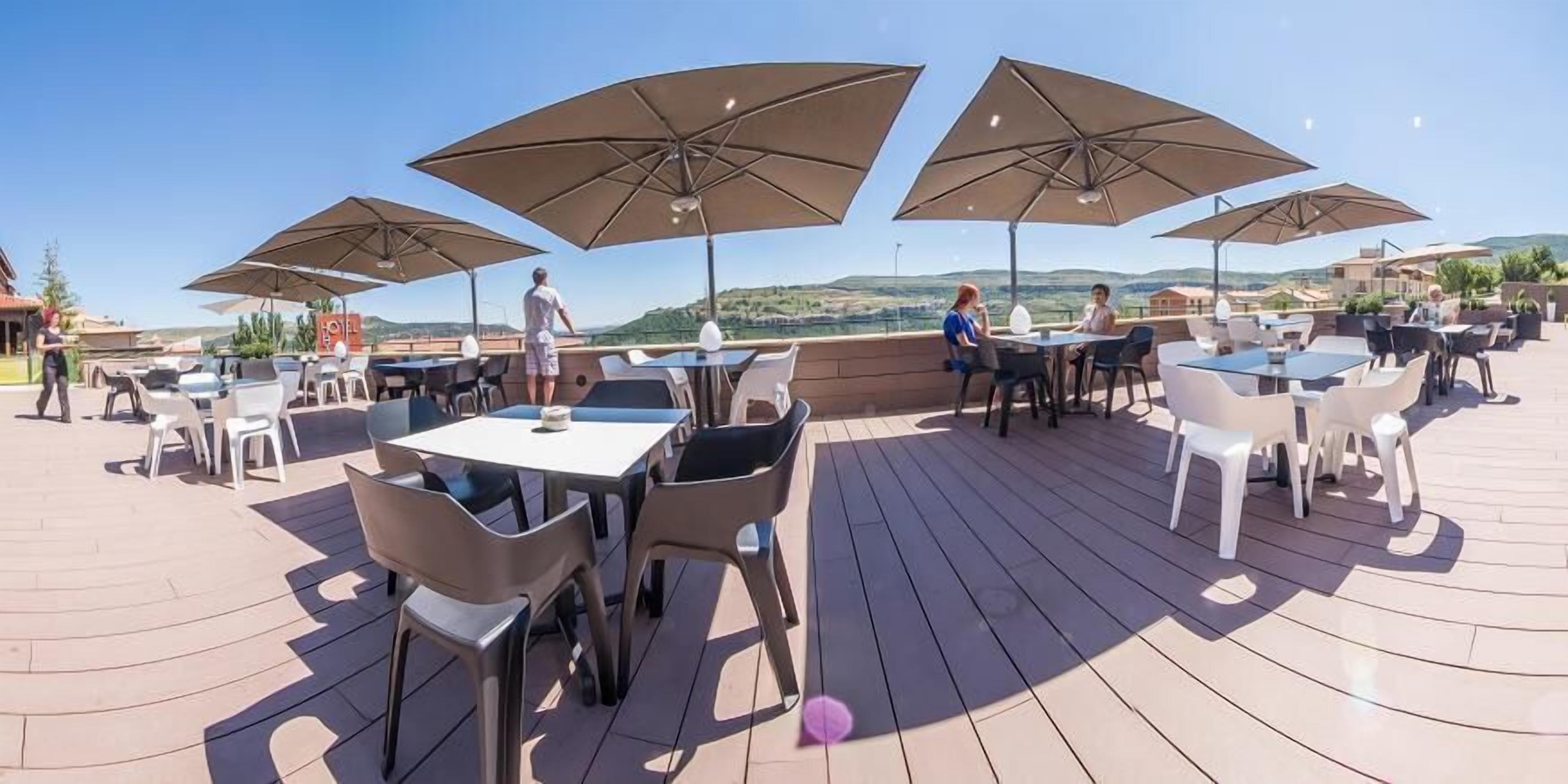 Un patio exterior con mesas y sillas, sombrillas grandes y vistas al paisaje.