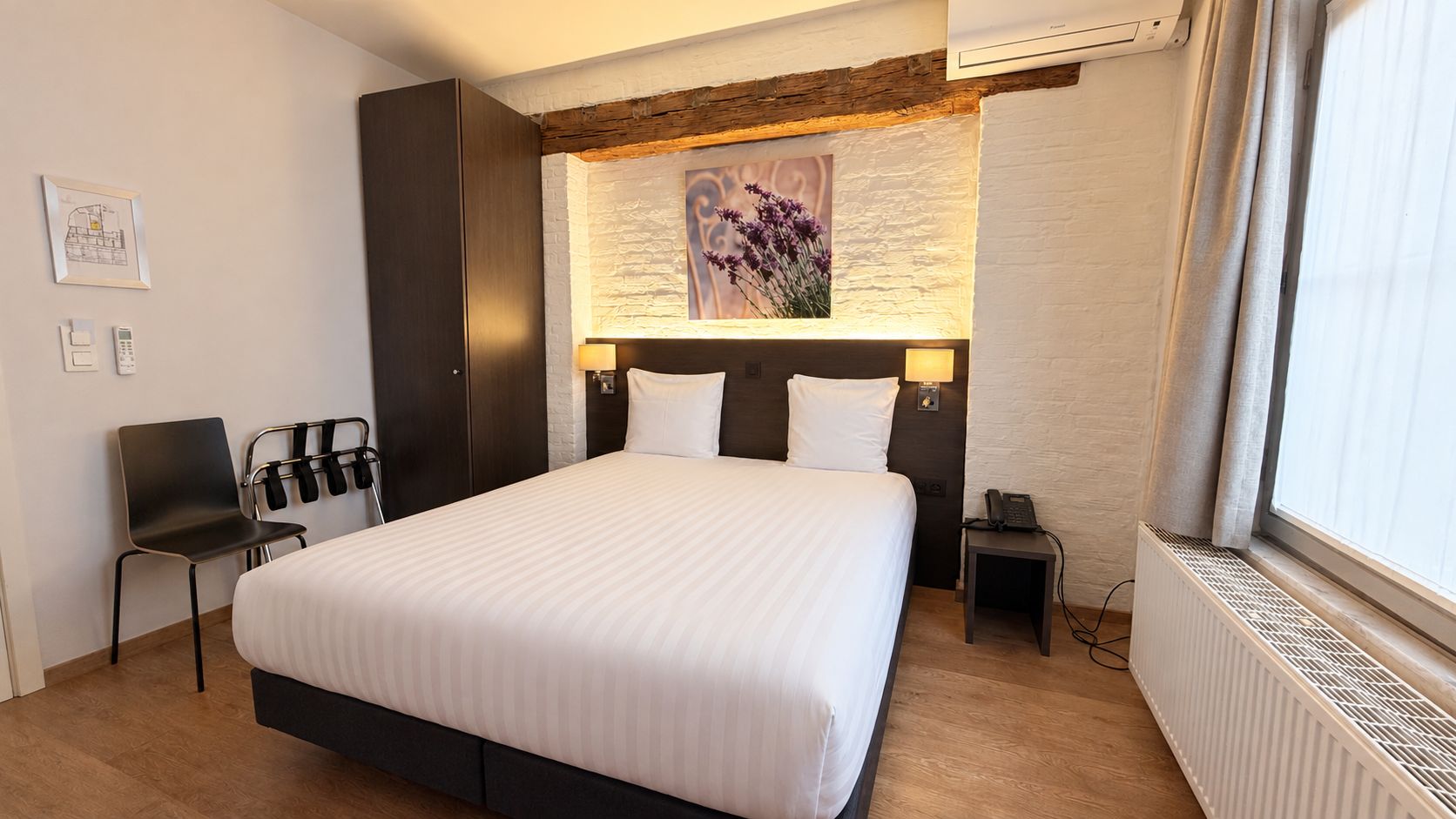 A hotel room with a large bed, a chair, a nightstand with a phone, a wall-mounted lamp, and a window with curtains.
