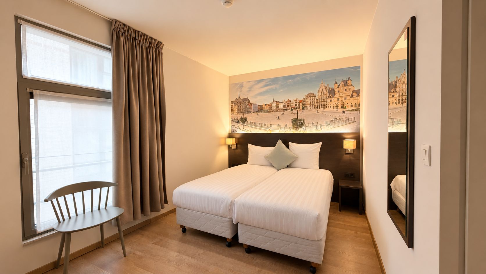 A hotel room with a bed, a chair, a window with curtains, a painting of a cityscape, and a mirror.