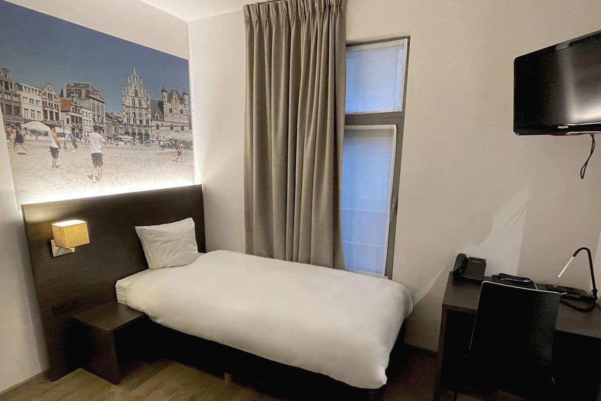 A hotel room with a bed, a desk with a chair, a TV mounted on the wall, and a window with curtains.
