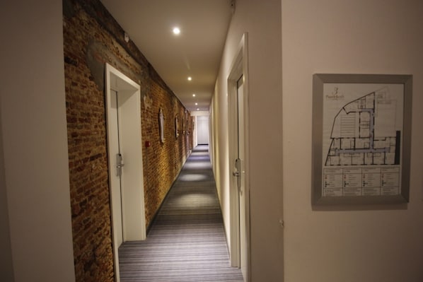 A corridor with a brick wall on one side and a white wall on the other, leading to a door at the end. A floor plan is displayed on the white wall.