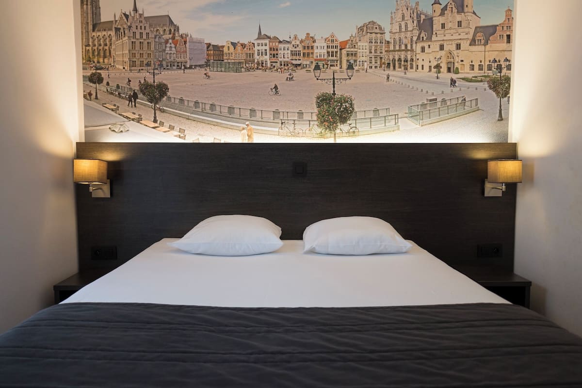 A hotel room with a large bed, two white pillows, a dark headboard, and two wall-mounted lamps.
