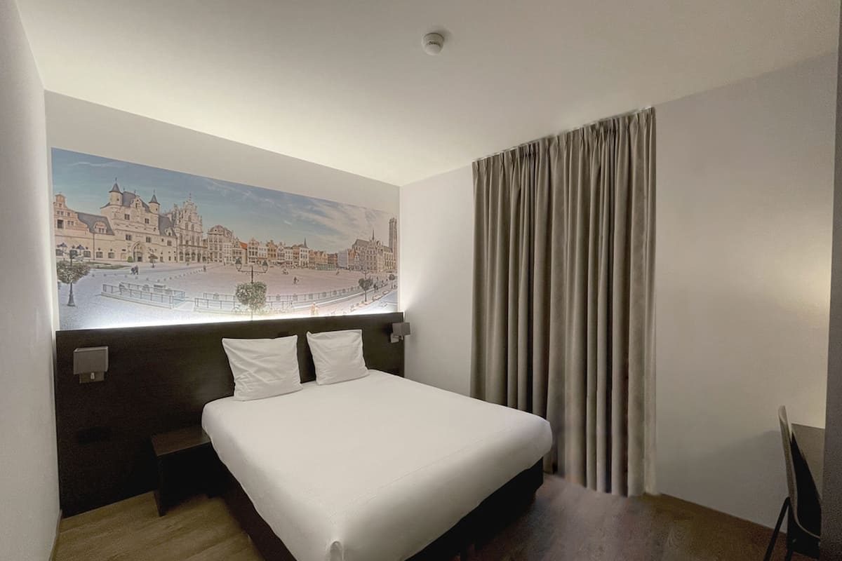 A hotel room with a bed, a headboard, a wall-mounted artwork of a cityscape, a window with curtains, and a wooden floor.