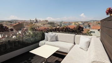 Terraço no telhado com um sofá branco, uma mesa pequena e guarda-corpo de vidro com vista para a paisagem urbana.