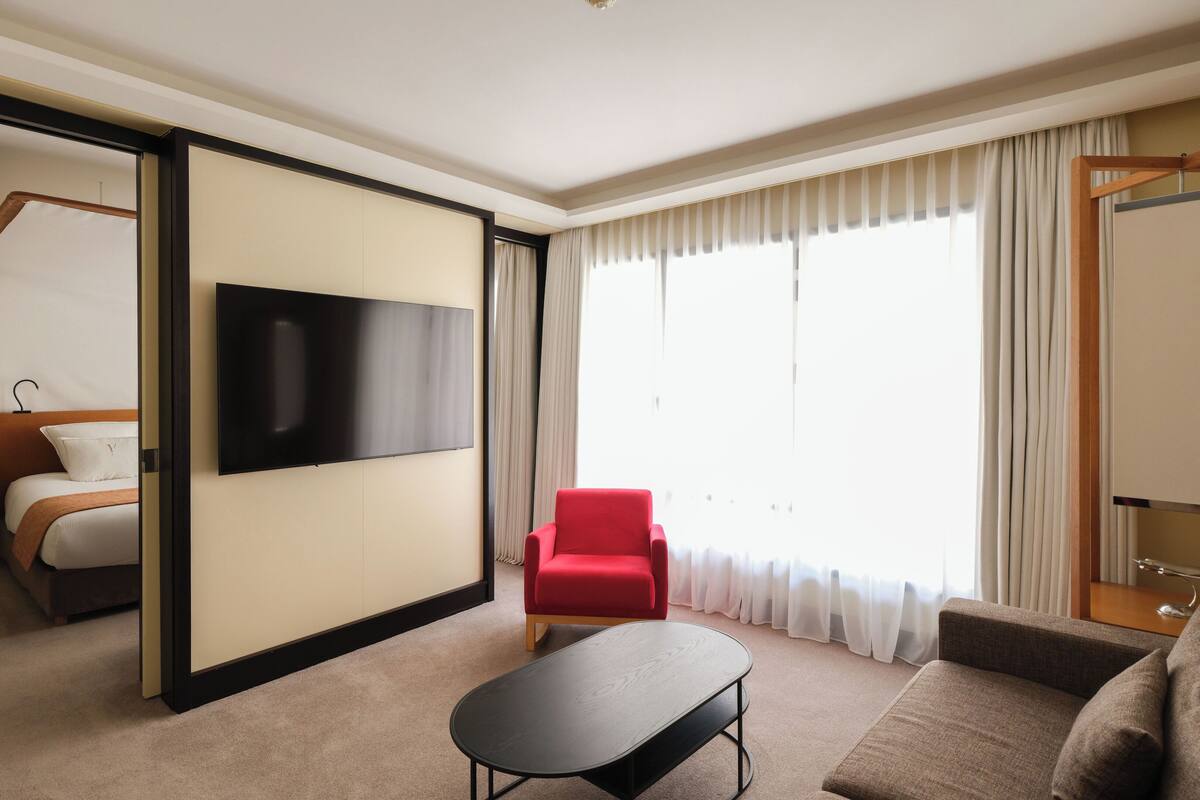 Habitación de hotel con un televisor de pantalla plana grande, un sillón rojo, un sofá y una pequeña mesa.