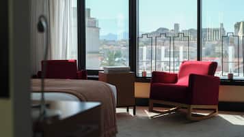 A hotel room with a large window, a red armchair, a small table, and a bed.