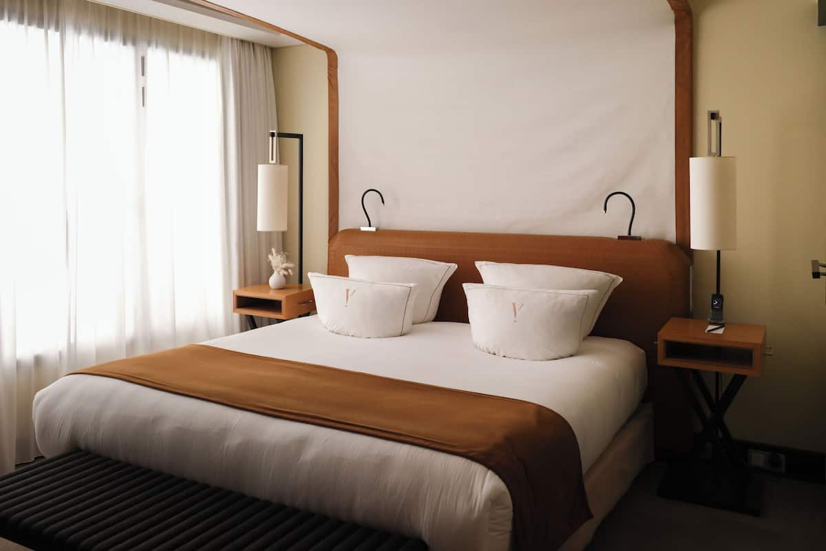 Habitación de hotel con una cama grande, dos mesitas de noche, un taburete y una ventana con cortinas.