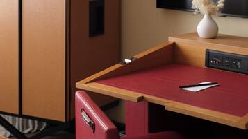 Une chambre d’hôtel compacte, dotée d’un bureau en bois, d’une chaise rouge, d’un mini-bar, d’un téléviseur à écran plat et d’un vase contenant des fleurs séchées, posé sur le bureau.