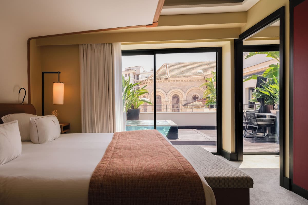 Habitación de hotel con una cama grande, una mesita de noche con lámpara, y vista a un patio con piscina y mobiliario exterior.