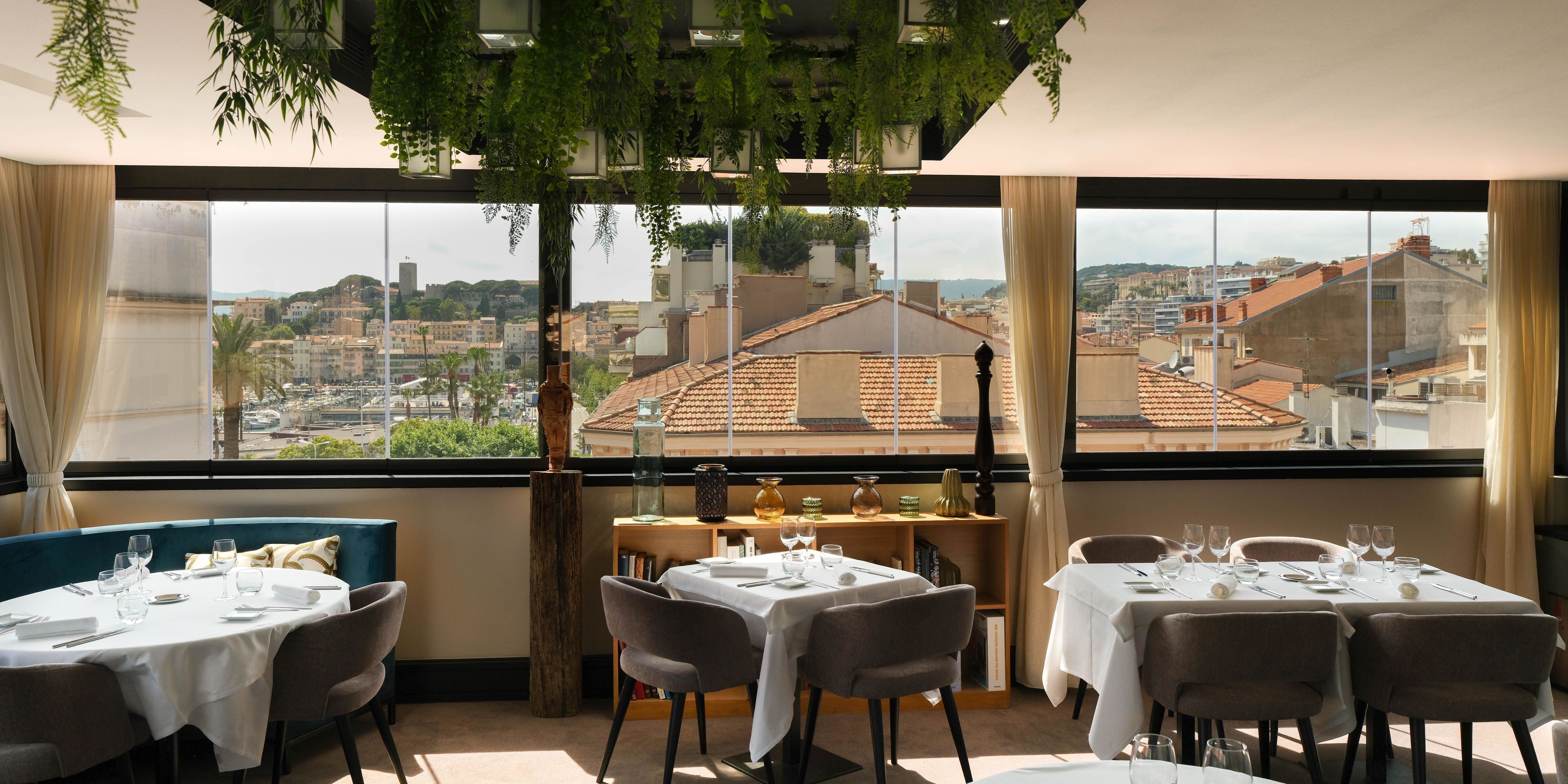 A restaurant with tables set for dining, a view of a cityscape through large windows, and indoor plants.