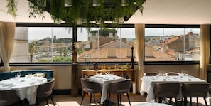 Un restaurante con mesas dispuestas para comer, vistas de un paisaje urbano a través de amplios ventanales y plantas de interior.