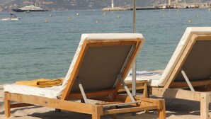Two wooden lounge chairs with white cushions and a white umbrella, positioned on a sandy beach with a view of the sea and a lighthouse in the background. - Five Seas by Inwood Hotels (Cannes)