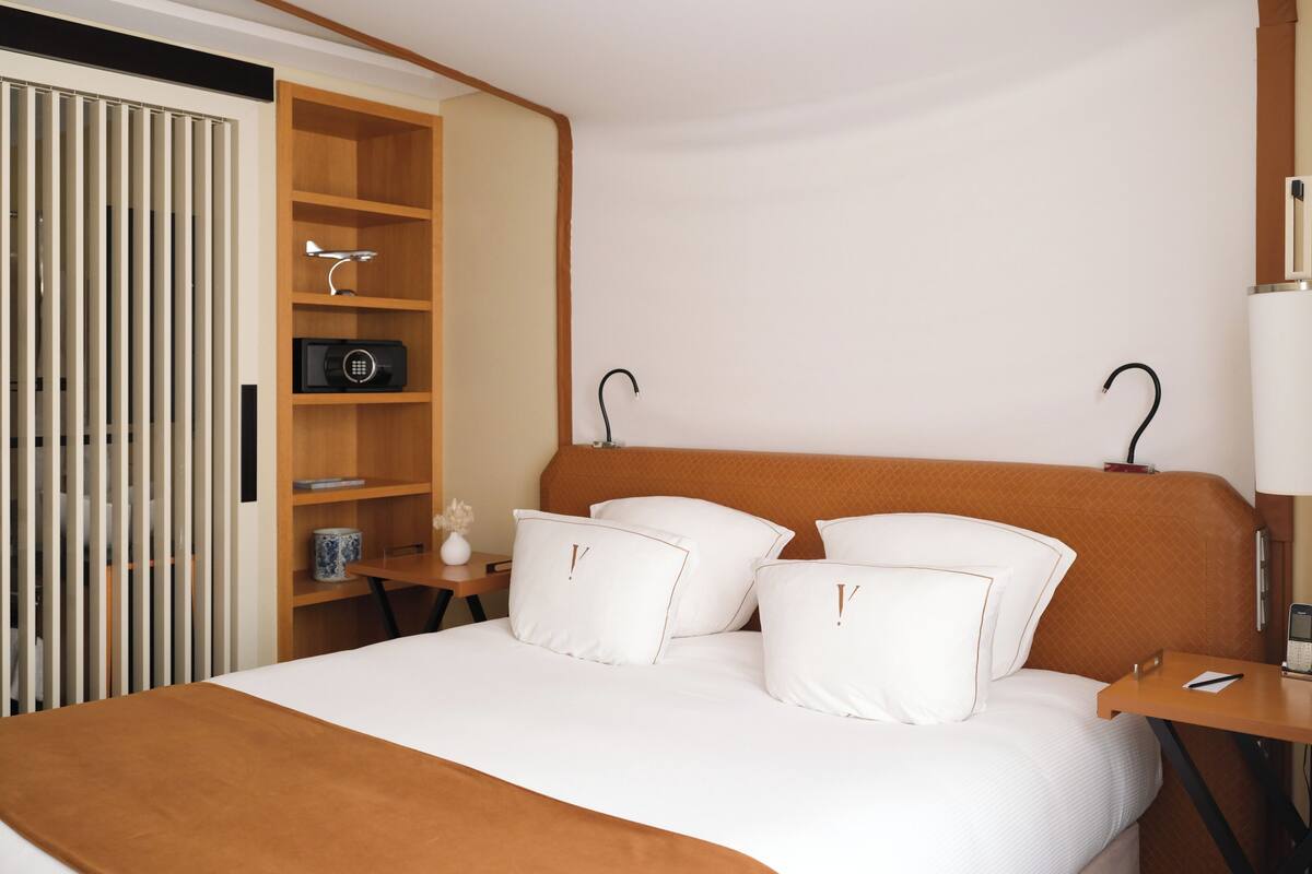 Una habitación de hotel con cama, mesita de noche, lámpara, estante con radio y armario.