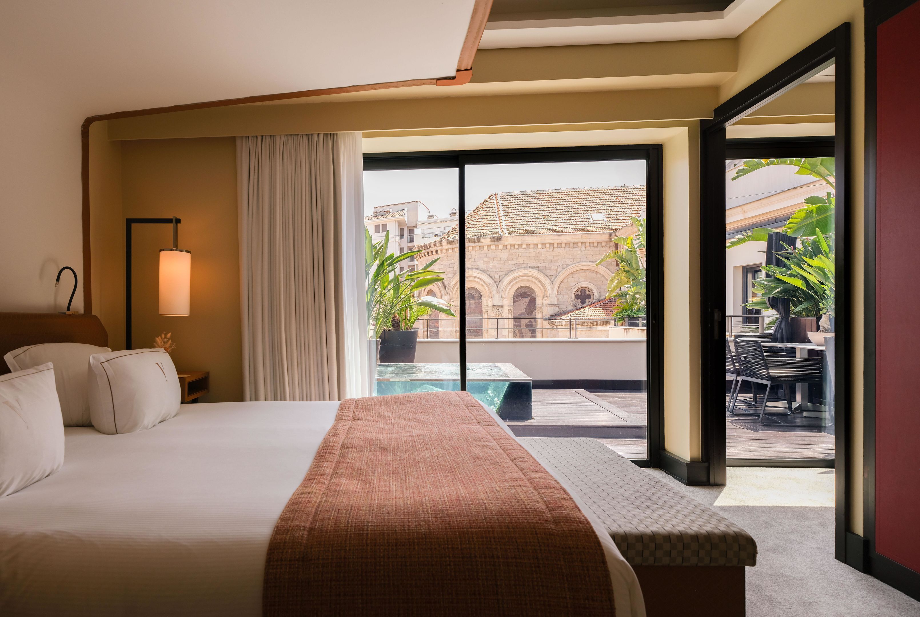 Une chambre d’hôtel avec un lit, une table de chevet, une lampe, une fenêtre avec des rideaux et une vue sur un immeuble et la verdure environnante.