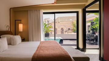 A hotel room with a bed, a nightstand, a lamp, a window with curtains, and a view of a building and greenery outside.