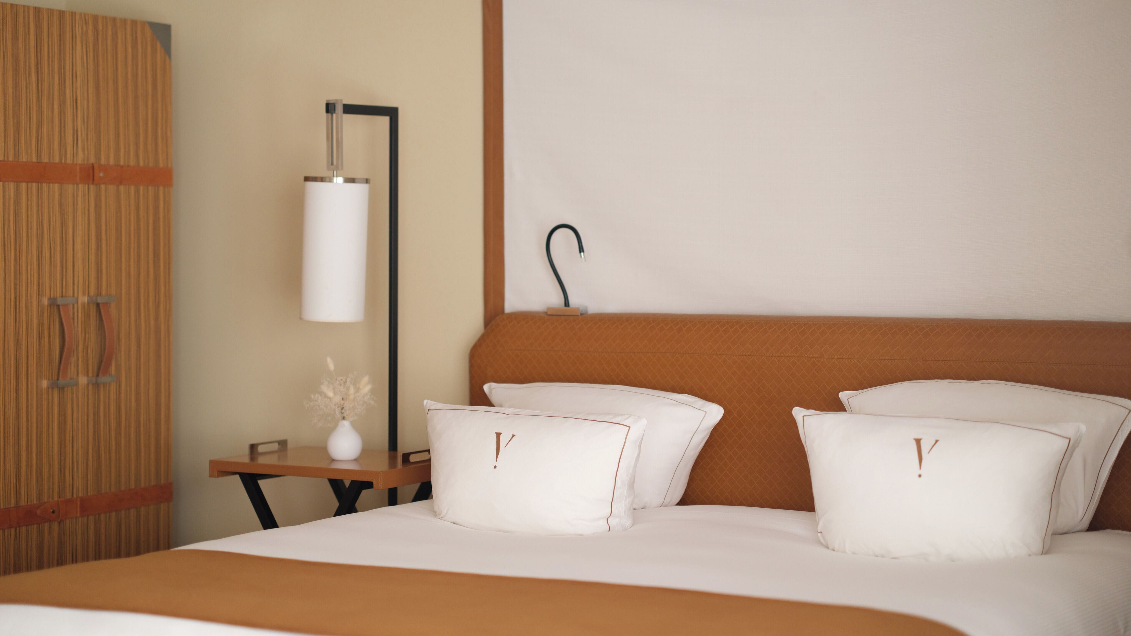 Habitación de hotel con una cama, dos almohadas, una mesita de noche con un florero y una lámpara de pared.