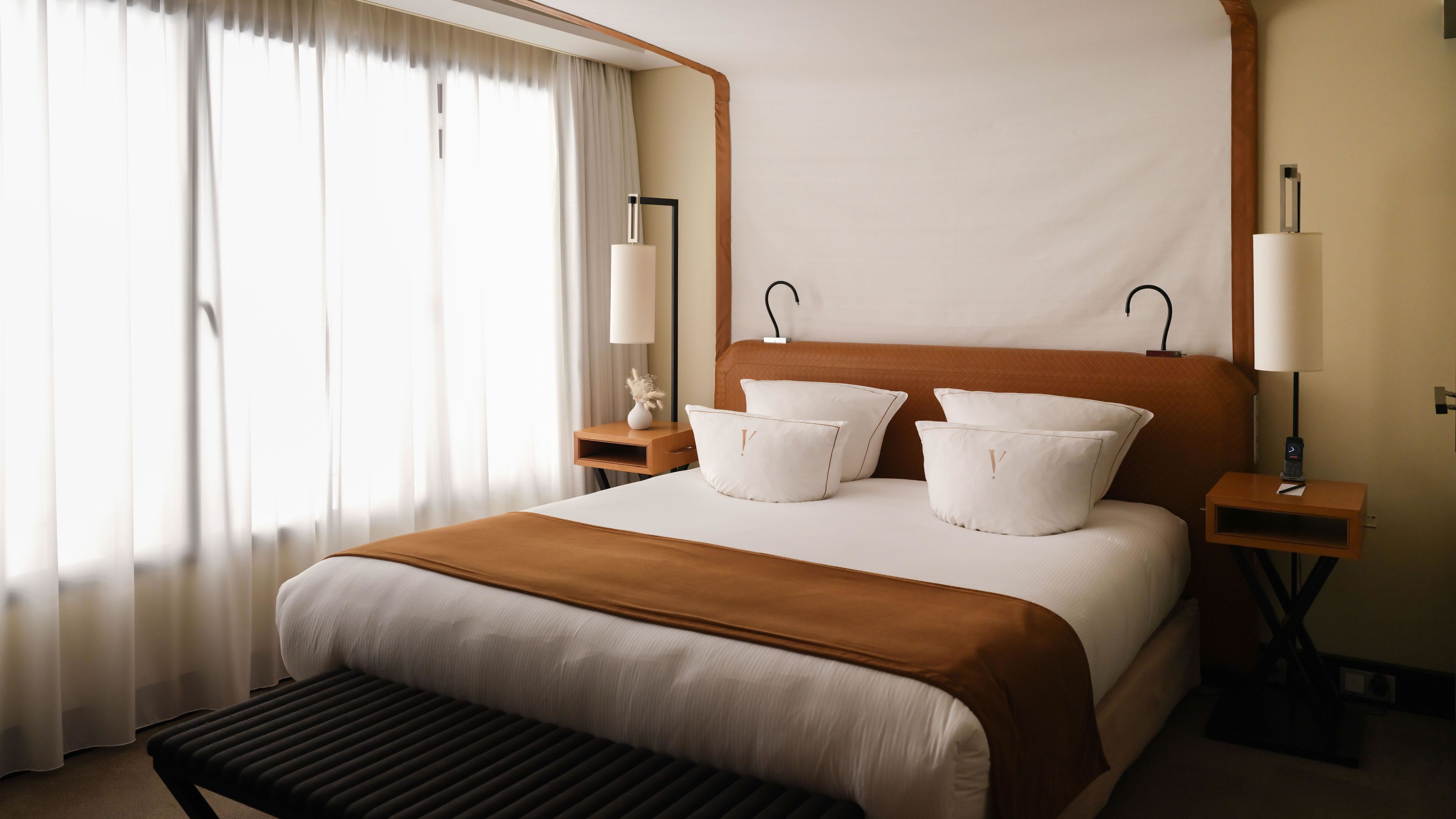 Habitación de hotel con una cama grande, dos mesitas de noche, un banco y una ventana con cortinas.