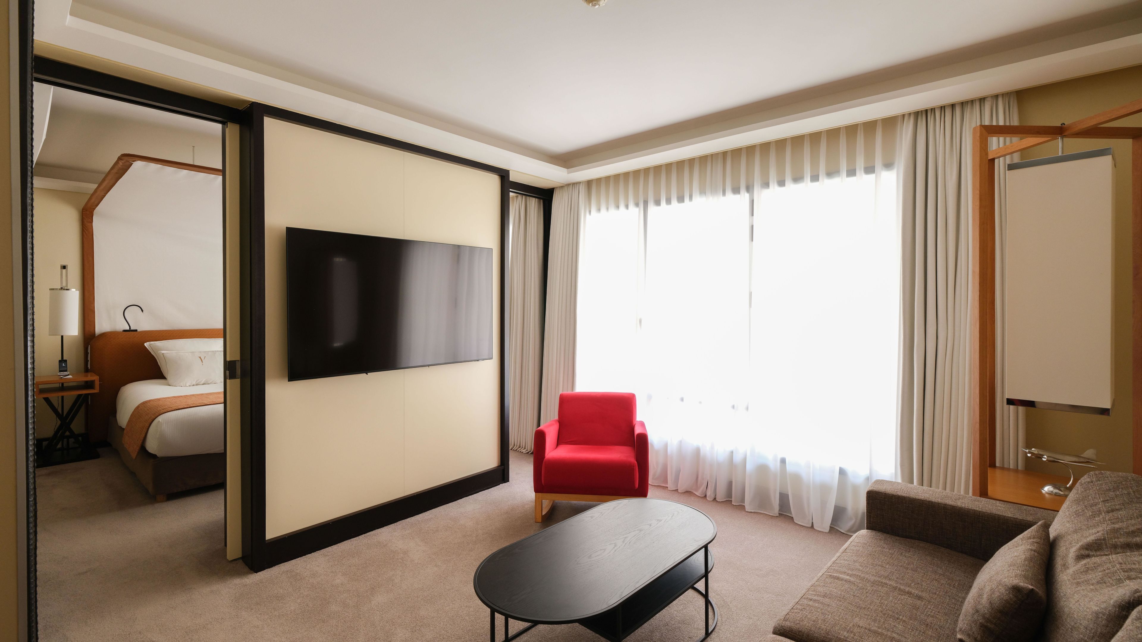 Ein Hotelzimmer mit einem Bett, einem Fernseher, einem roten Sessel, einer Couch und einem großen Fenster mit Vorhängen.