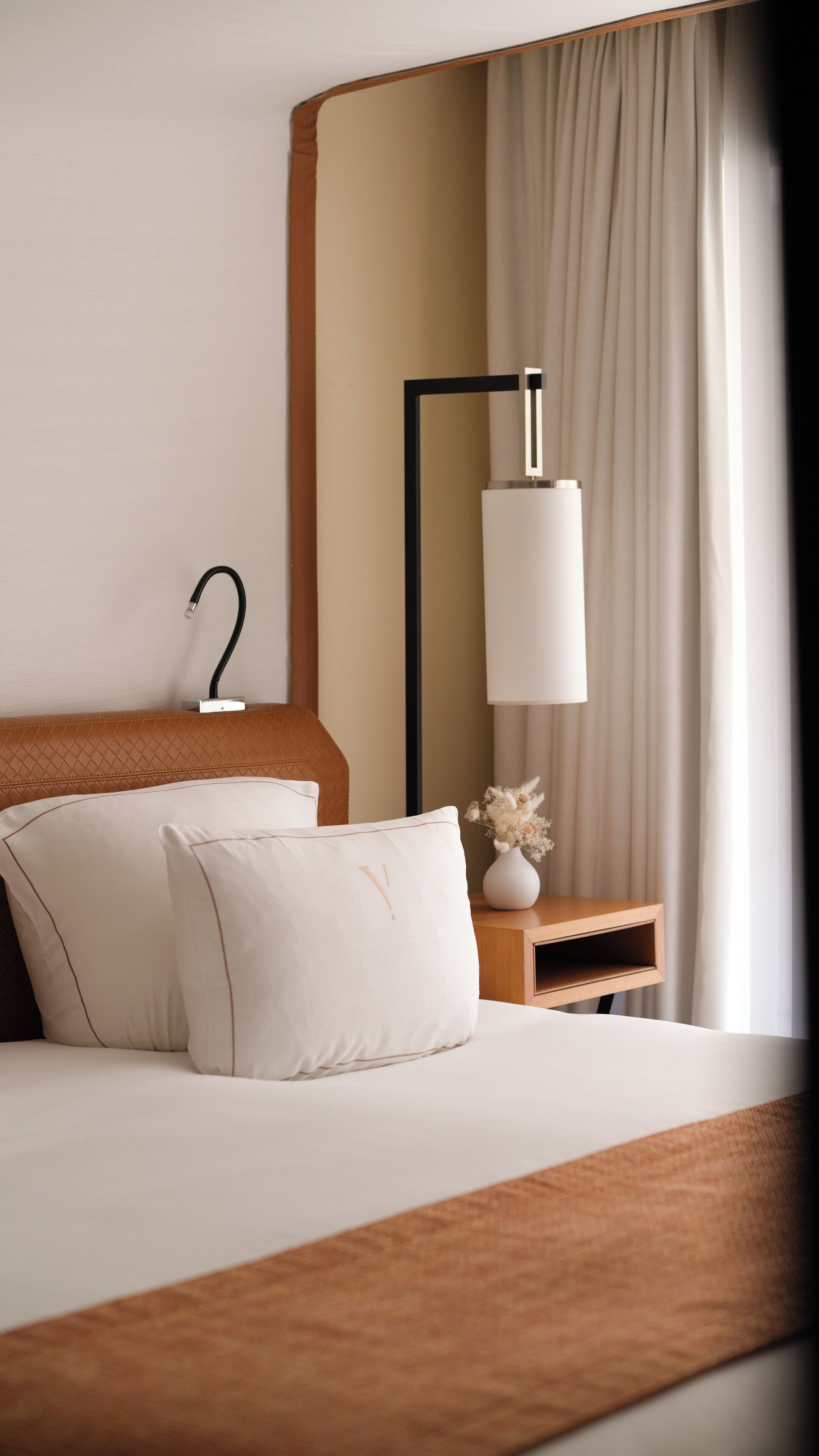 Habitación de hotel ordenada con una cama, una mesita de noche con una lámpara y un florero con flores secas.