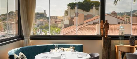 Un comedor con una mesa dispuesta para dos personas, vistas de la ciudad a través de amplios ventanales y una estantería con libros.
