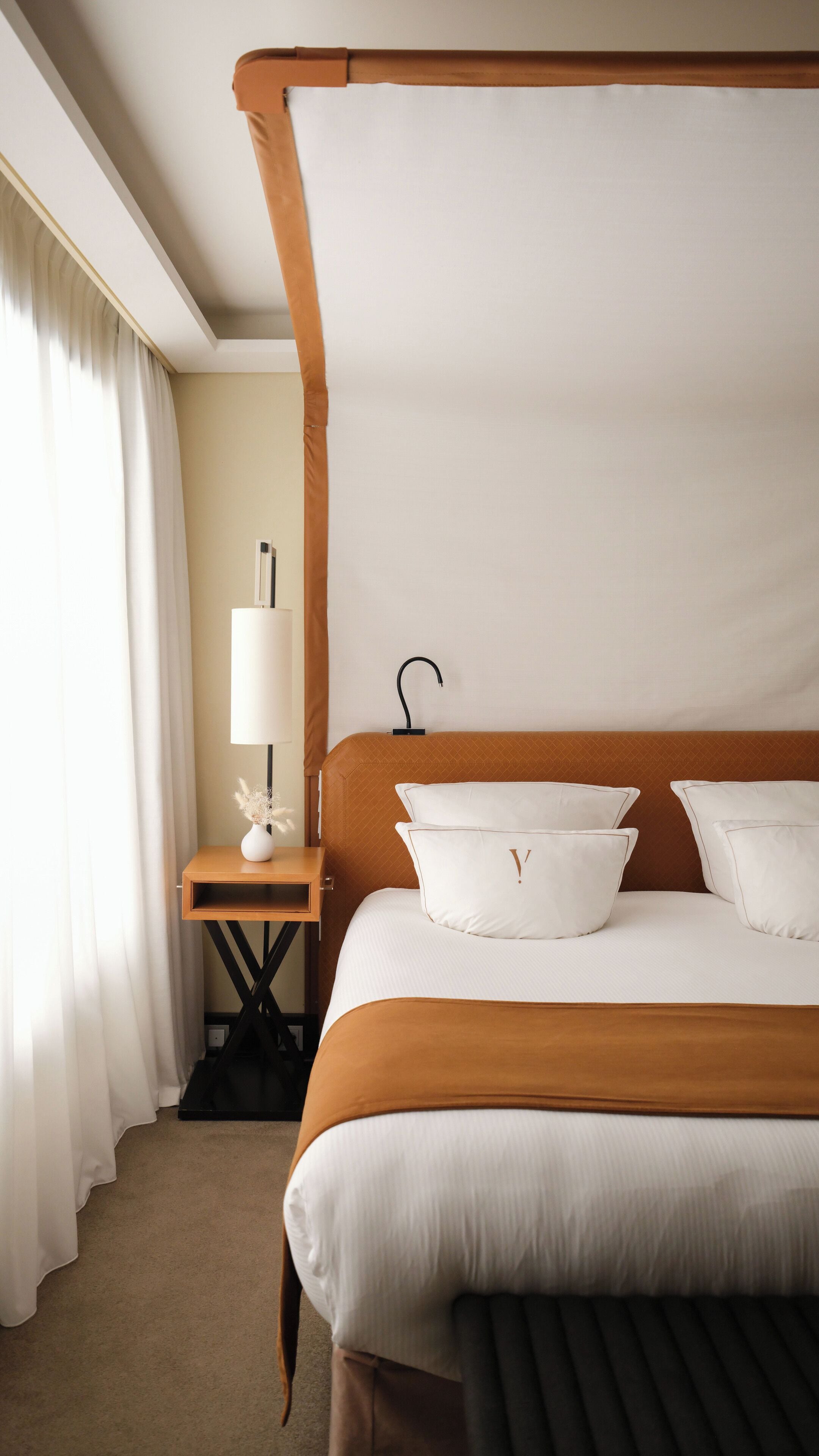 A hotel room with a four-poster bed, a bedside table with a lamp, and a vase with dried flowers.
