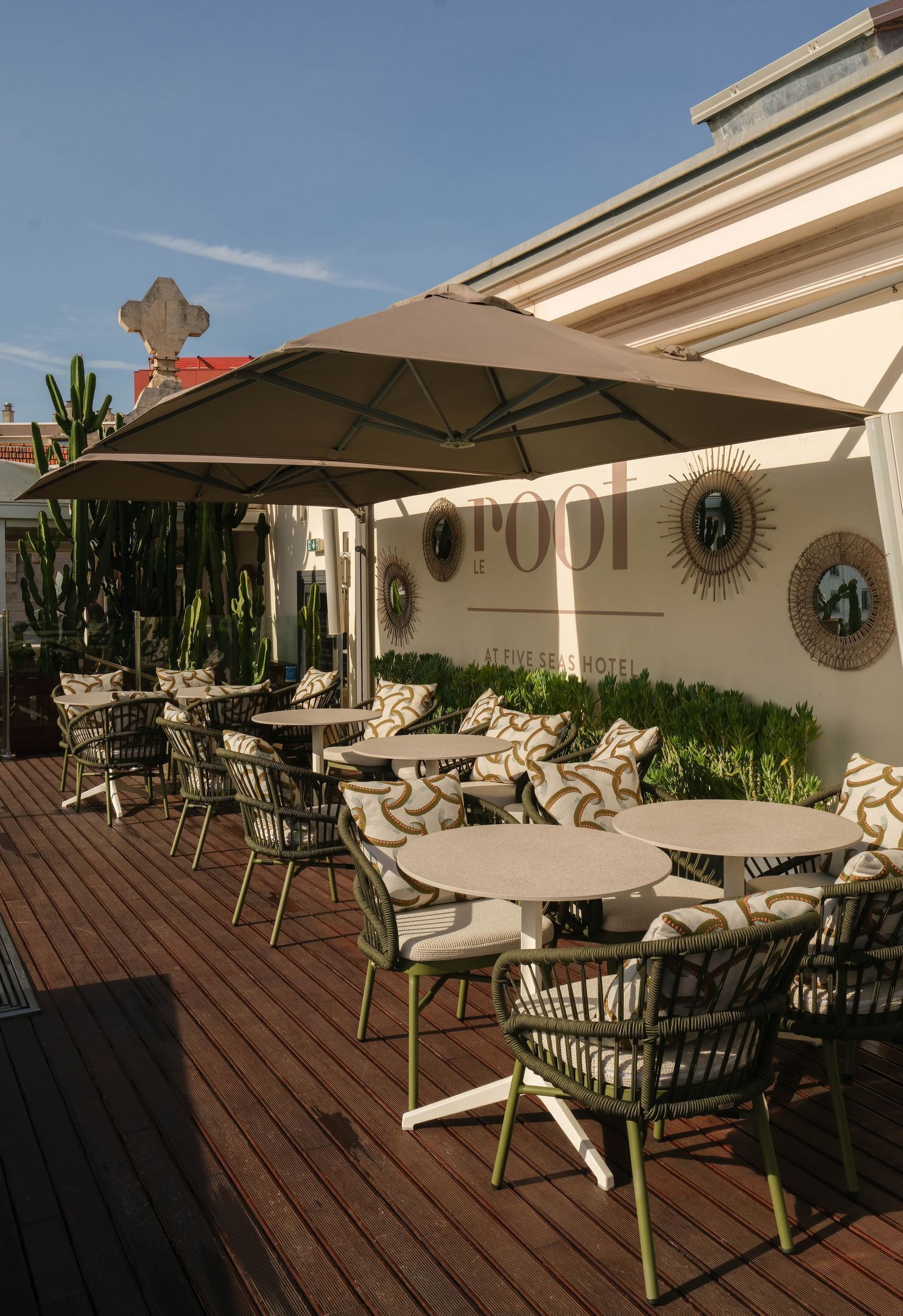An outdoor seating area with round tables and wicker chairs, a large umbrella, and a wall with a decorative mirror and wall art.