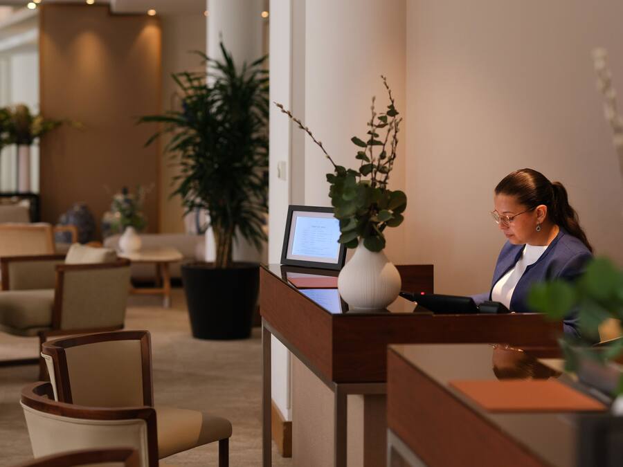 Taulell de recepció amb un ordinador, cadires i plantes en testos en un vestíbul d'hotel.