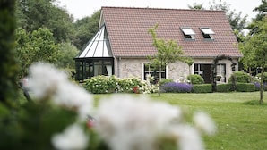 Una casa con techo inclinado, un invernadero y un jardín bien cuidado con diversas plantas y flores.