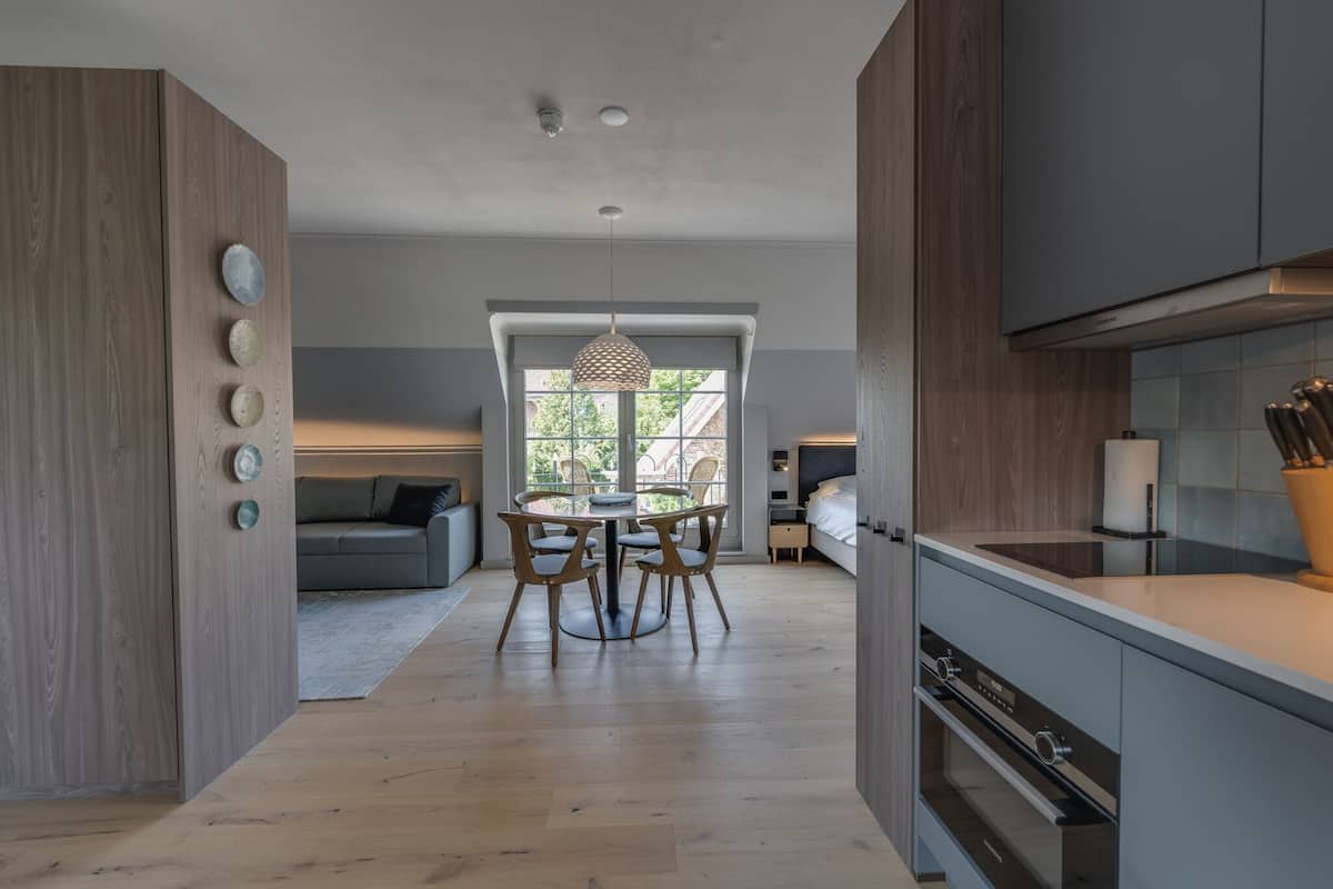 A modern kitchen with wooden cabinets, a dining area with a round table and chairs, and a view of a living room with a sofa and a window.