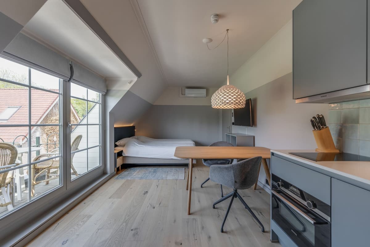 A modern, compact bedroom with a large window, a wooden desk, and a kitchenette.