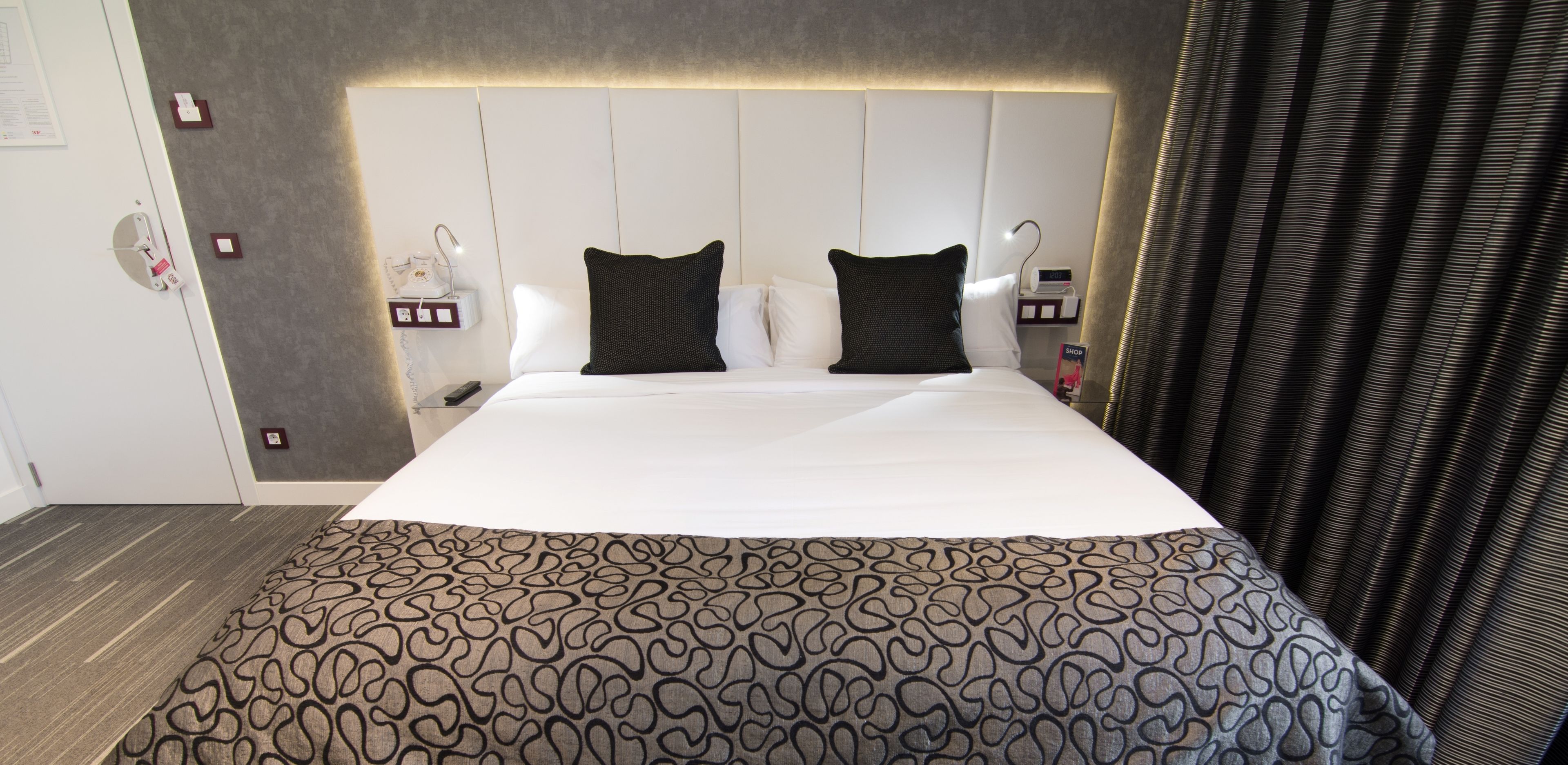 Habitación de hotel con una cama grande, dos almohadas, una mesita de noche con lámpara, un enchufe y un reloj de pared.
