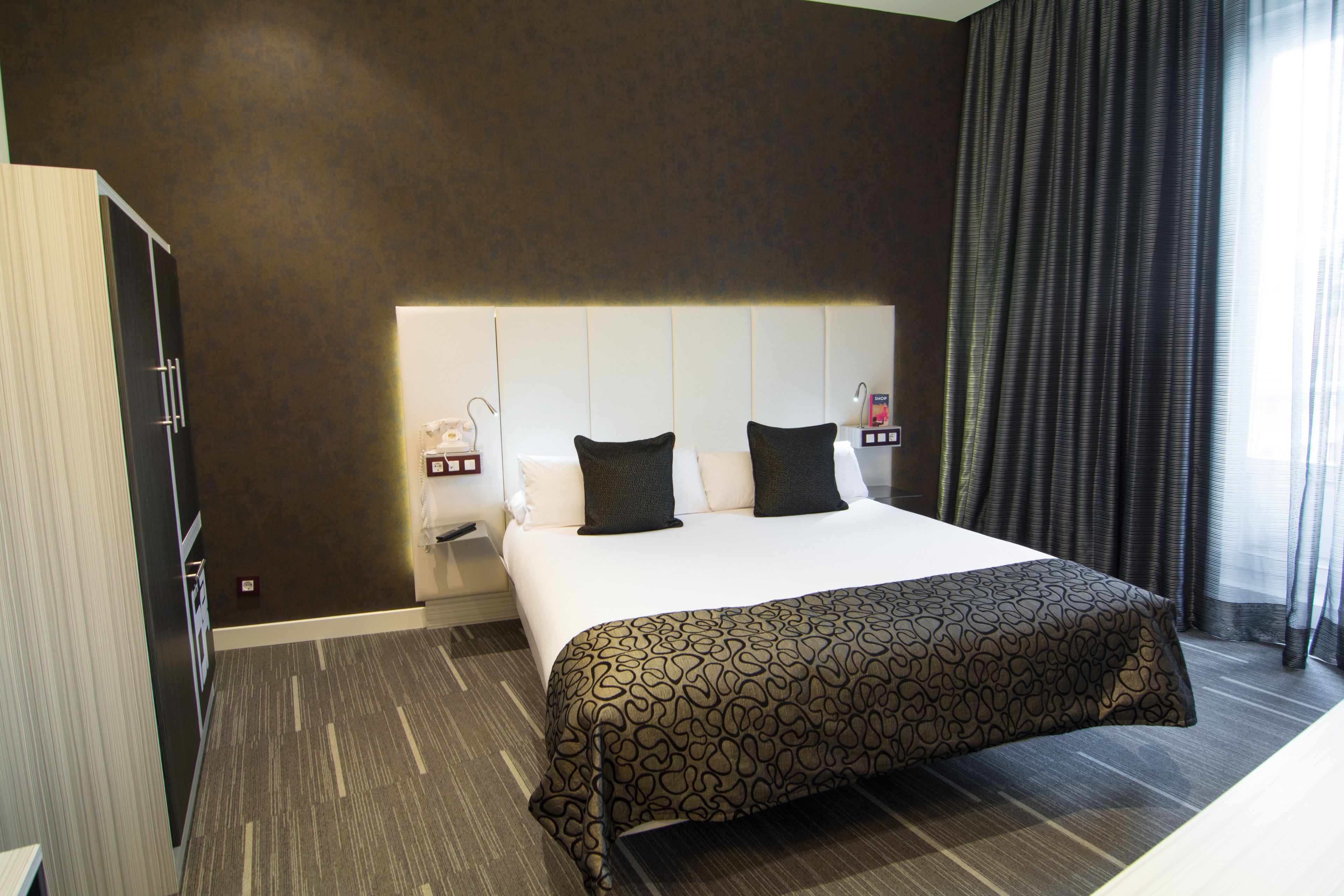 A hotel room with a large bed, a nightstand, a wardrobe, and a window with curtains.