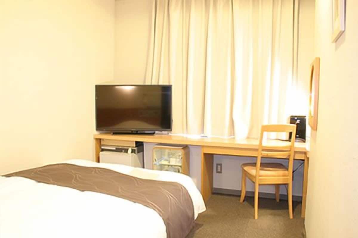 A hotel room with a bed, a desk with a television, a chair, and a small table.