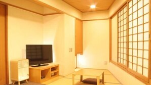 Een traditionele Japanse kamer met tatami vloerbedekking, een televisie op een houten standaard, een klein kastje en een schuifdeur met een rasterpatroon.